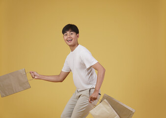 portrait of happy young asian man dressed casually holding shopping bags isolated on yellow background