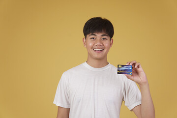 portrait of happy young asian man holding credit card for shopping payment isolated on yellow background