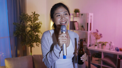 happy asian chinese woman singing karaoke and drinking bottle of alcohol while enjoy free time alone at home living room.