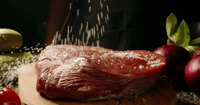 Chef cooks beef steak, sprinkling with salt on a background of professional kitchen, slow motion.