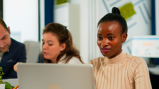 Portrait Of Delighted African Business Woman Reading Good News On Laptop Sitting At Desk In Busy Start Up Office While Diverse Team Analyzes Statistics Data. Multiethnic Team Working At New Project