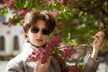 Portrait of a young woman in sunglasses near a lilac bush in a gray coat on a sunny spring day