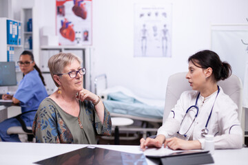 Fototapeta premium Senior woman showing to doctor sore throat during medical consultation sitting in hospital room. Elderly patient visiting doctor examining thyroid throat touching health at clinic, treatment medical