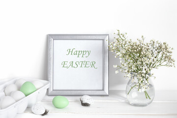 White and green Easter eggs, a mockup with a silver frame and spring flowers in a vase on a white wooden table