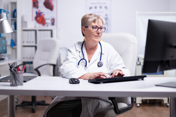 Fototapeta na wymiar Senior female doctor in lab coat, glasses and stethoscope over her neck typing on computer sitting in medical office using looking at desktop smiling while working in medical office