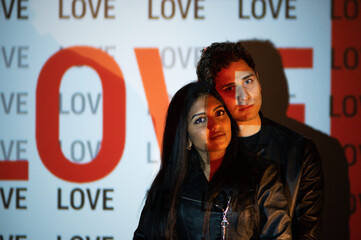 Loving Indian and Caucasian couple with black jacket looking at the camera. Lovers in dark studio illuminated by light of projector with word love.