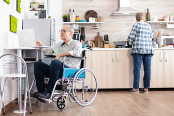 Disabled senior man in wheelchair working from home at laptop in kitchen while wife is cooking...