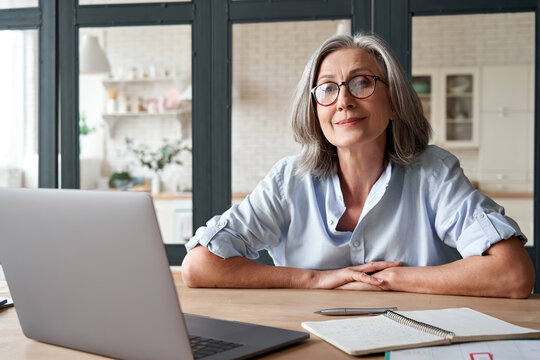 Smiling Stylish Mature Middle Aged Woman Sits At Desk With Laptop, Portrait. Happy Older Senior Businesswoman, 60s Grey-haired Lady Wearing Glasses Looking At Camera Sitting At Office Table. Headshot.