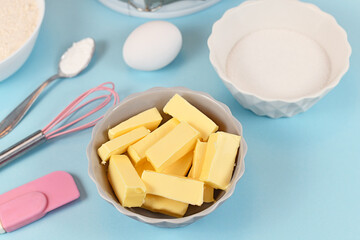 Butter cut in pieces for preparing dough for shortcrust cake base with sugar, egg, baking bowder and baking utensils on blue background