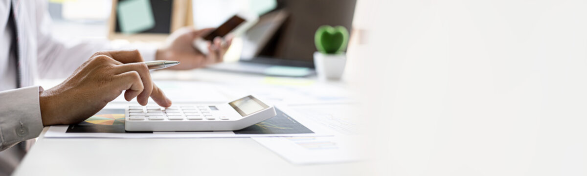 Businessman Is Using A Calculator To Calculate Company Financial Figures From Earnings Papers, A Businessman Sitting In His Office Where The Company Financial Chart Is Placed. Banner With Copy Space.