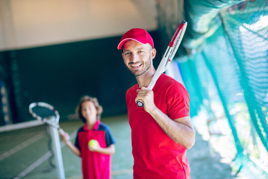 Young bearded man in a red cap holding a tennis racket and talking to a boy