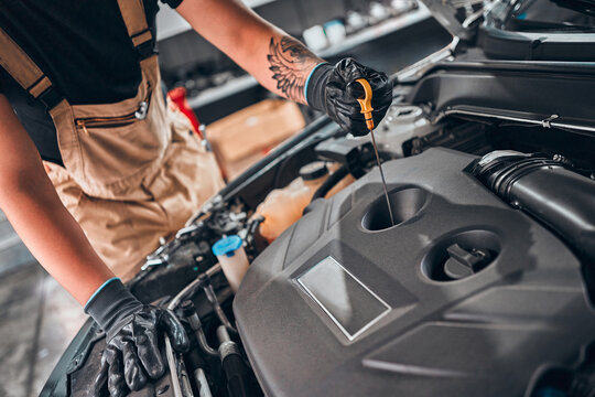 Concept of male car mechanic holding dipstick checking level oil on the car engine, for repair and maintenance services, wearing a black glove and beige garment overalls with front car engine.