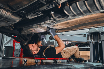 Young mechanic lying down and working under car at the garage.