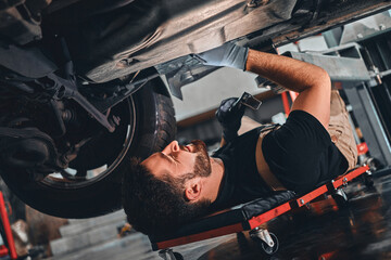 Bearded male technician in gloves lying on car creeper and using LED inspection lamp while working under automobile in garage.