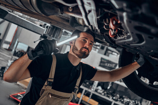 Portrait shot of a handsome mechanic working on a vehicle in a car service. Professional repairman is wearing gloves and using a lamp underneath the car.