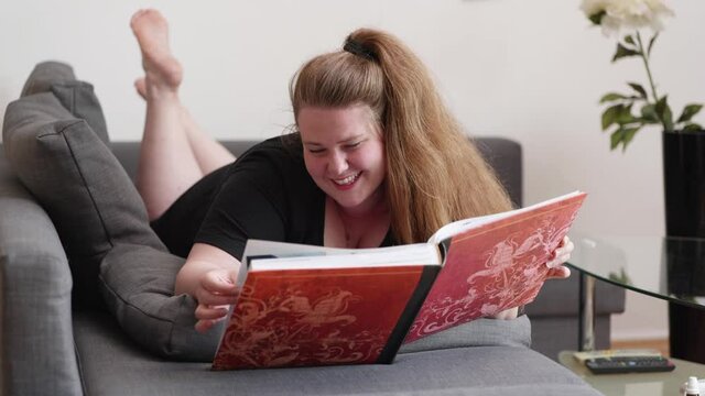[4k] authentic brunette woman lying on couch looking at a red picture book