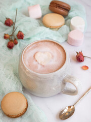 Fototapeta na wymiar cup of hot chocolate with heart shape marshmallow and pieces of varieties French macarons with dried roses in background. Warming valentine's drink 