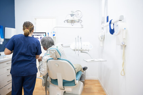 View from behind of Female dentist and mature patient sitting in chair in dental clinic treatment room while showing her the screen with images of his treatments.