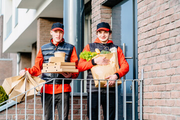 Two delivery men holding pizza box, take-out food containers, coffee cups in holder and paper bag, boxes. Building or apartment house on background. Delivery teamwork concept.