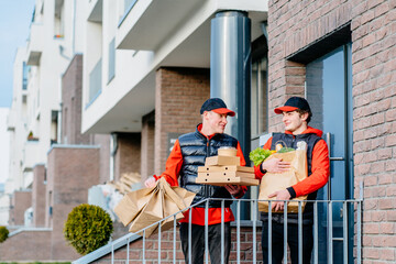 Two delivery men holding pizza box, take-out food containers, coffee cups in holder and paper bag,...