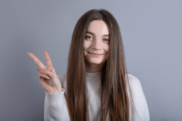 Portrait of a young attractive girl with dark long hair sgesture V sign for victory or peace sign on a gray studio background. Place for text