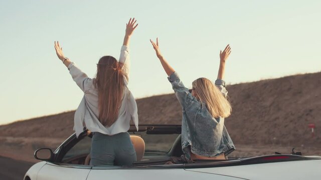 Two lifestyle freedom women with long hair standing in convertible and raising their hands up, background of hill and sky. Woman friends ride standing in back seat of convertible, rear view. Concept