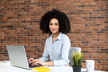 Young American businesswoman sitting at the laptop and looking at the camera, smiling and typing, interrupted from work, best worker, drinking coffee, love her work,beautiful neat girl in formal shirt