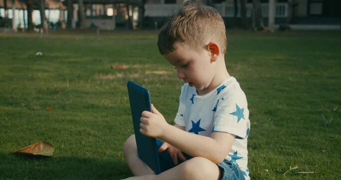 Curious cute baby boy preschool child using a digital tablet technology device looks telephone sitting on the street on the grass in the park. Kid plays in the tablet.