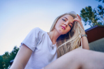 Charming teenage girl in a white T-shirt against a blue sky sits in a park and straightens her hair. View from below.