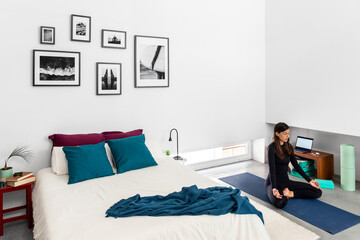 Young woman practicing yoga and meditation in lotus pose in bedroom with minimalist style interior