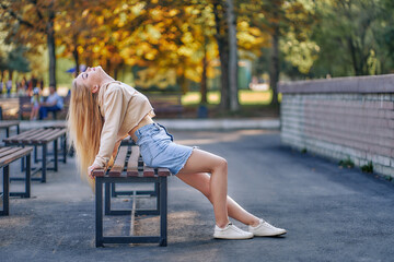 A cute woman in a denim skirt and a blouse is leaning back sitting on the autumn playground of the park.