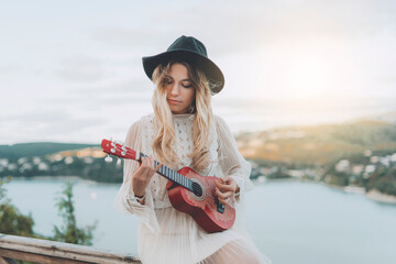 A girl with blond hair in a white boho dress and a hat is playing a ukulele on the observation deck near a turquoise lake