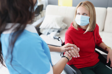 Woman in wheelchair in protective medical mask gives hands to doctor. Coronavirus and disability...