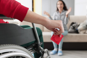 Person sitting in wheelchair holds red condom in his hand in front of woman sitting. Safe and...