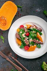 Baked pumpkin salad, spinach, tomato and beef in a white plate on a stone background