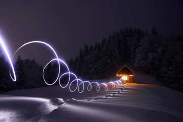 Winter night. House with light. The wide trail leads to the snowy old wooden hut. Landscape of high mountains and forests. Lawn covered with snow. Location place Carpathian, Ukraine, Europe.