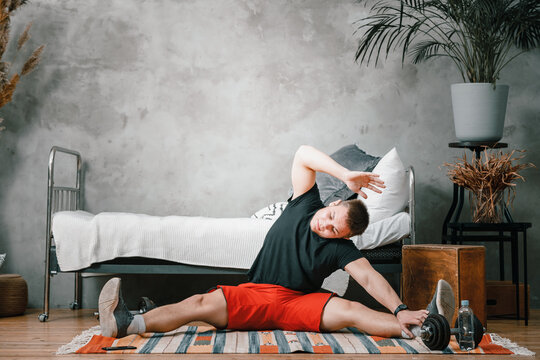 A young man goes in for sports at home, online workout . The athlete  stretching , meditating, sitting on a twine  in the bedroom, in the background there is a bed,  a carpet.