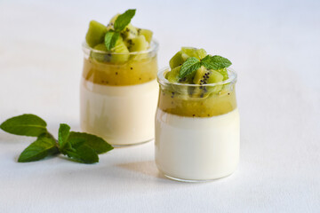Italian dessert panna cotta in glass jars with kiwi fruit jelly decorated with pieces of fresh kiwi and mint leaf on white background. Copy space