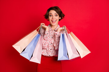 Portrait of lovely amazed cheerful brown-haired girl carrying buyings new things having fun isolated over bright red color background