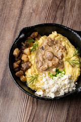 Banosh - a traditional dish in the Western Ukraine made of cornmeal. Served in a black pan with cracklings, mushrooms and hutsul cheese bryndza. Close-up top view isolated on a wooden background.