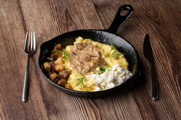 Banosh - a traditional dish in the Western Ukraine made of cornmeal. Served in a black pan with cracklings, mushrooms and hutsul cheese bryndza. Close-up isolated on a wooden background with cutlery.