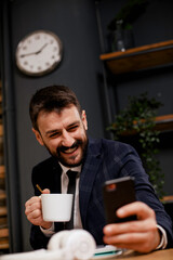 Young businessman using phone and drinking coffee in his office. Handsome man on coffee break.