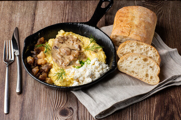Banosh - a ukrainian dish made of cornmeal. Served in black pan with cracklings, mushrooms and hutsul cheese. Close-up isolated on a wooden background with cutlery, sliced bread and grey linen napkin.