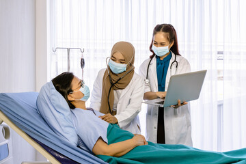 Team of doctors visiting and taking care of senior female patient in bed, checking heartbeat with stethoscope, and using laptop computer.