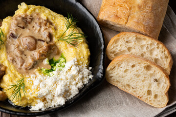 Banosh - a traditional ukrainian dish made of cornmeal. Served in black pan with cracklings, mushrooms and hutsul cheese bryndza. Macro close-up on a grey linen napkin with sliced bread. Top view.