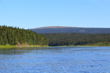 Panoramic photo of a lake with calm water with a kayak in the distance, with tall pine trees overgrown with dense forest with a towering mountain on the horizon and a blue sky