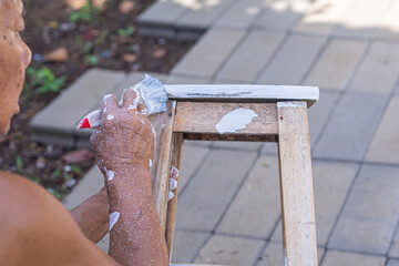 Senior man hand paints with brush an old wooden chair without a backrest. Hand of man. Old chairs...
