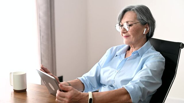 Charming senior businesswoman in glasses sitting at the desk, using a tablet for video chatting, an elder female English teacher is scrolling news online, looking at the photos and smiling