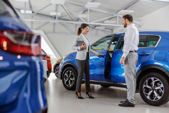 Smiling Friendly Female Car Seller With Tablet In Hands Opening The Car Door And Inviting Buyer To Sit And Try A New Car. Car Salon Interior.