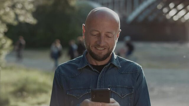 Bald and handsome man in his mid-forties with a stylish beard is standing in the park and is chatting with his friedns on a mobile. He is smiling wildly.